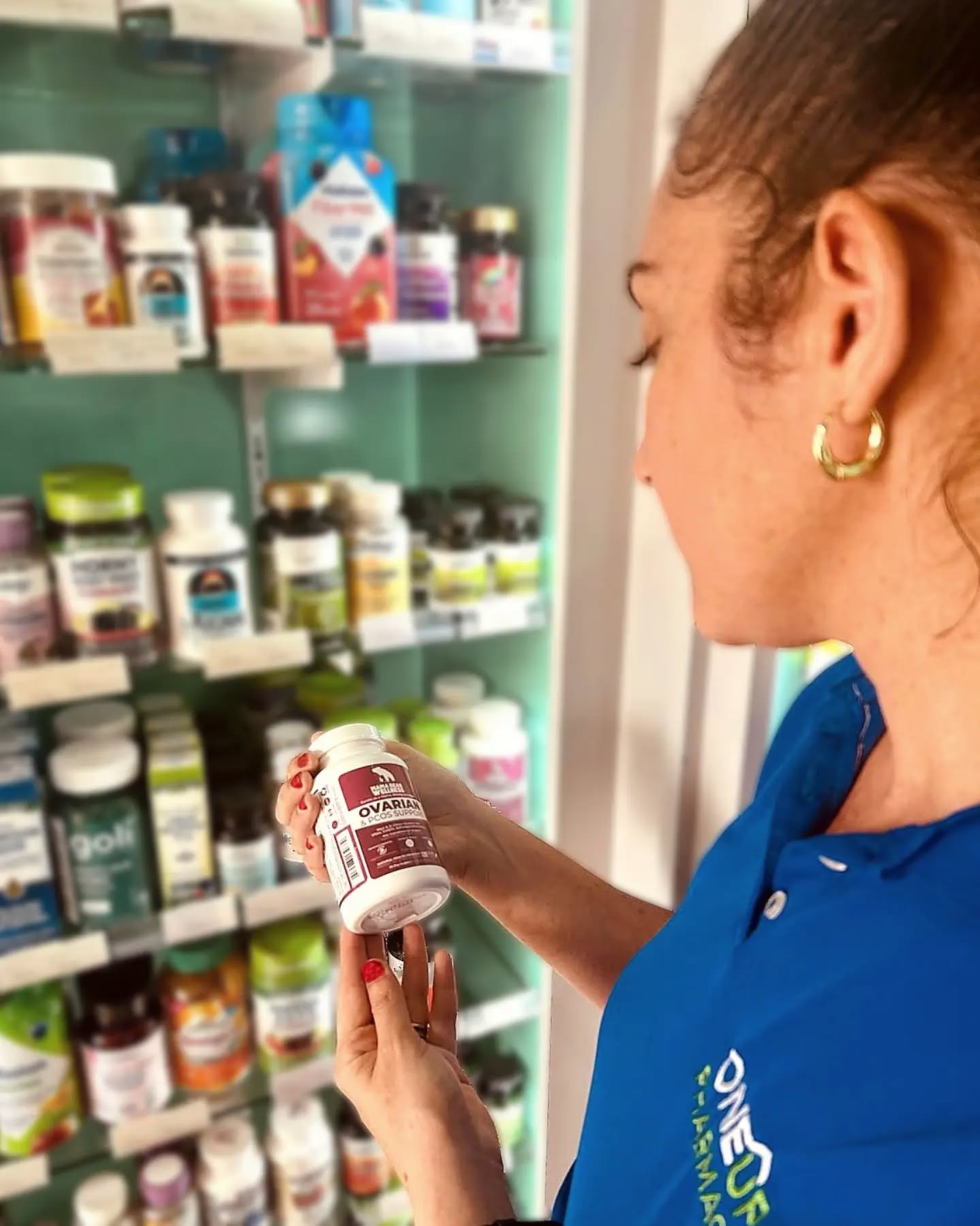 Pharmacist in a blue One Care Pharmacy polo examining a bottle of Mama Bear Ovarian & PCOS Support in front of stocked supplement shelves